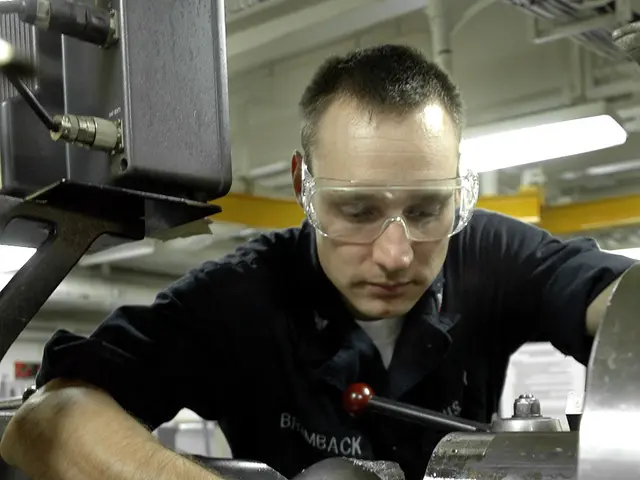 The image shows a machinist wearing glasses and working on a machine in a factory, illuminated by...