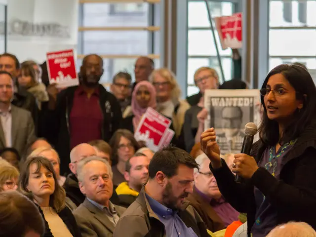 The image shows a woman wearing glasses and holding a microphone in front of a crowd of people,...
