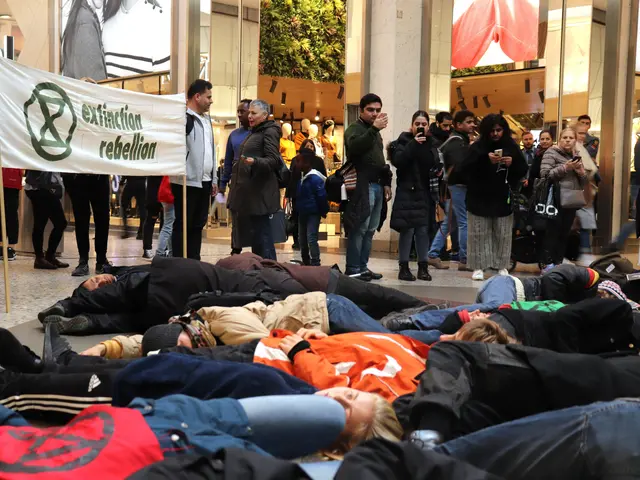 The image shows a group of people laying on the ground in front of a crowd of people, some of whom...