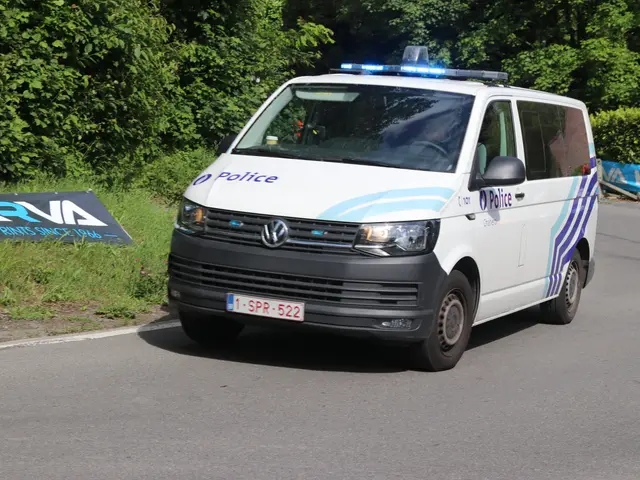 The image shows a police van driving down a road next to a forest. The van is white, blue and black...