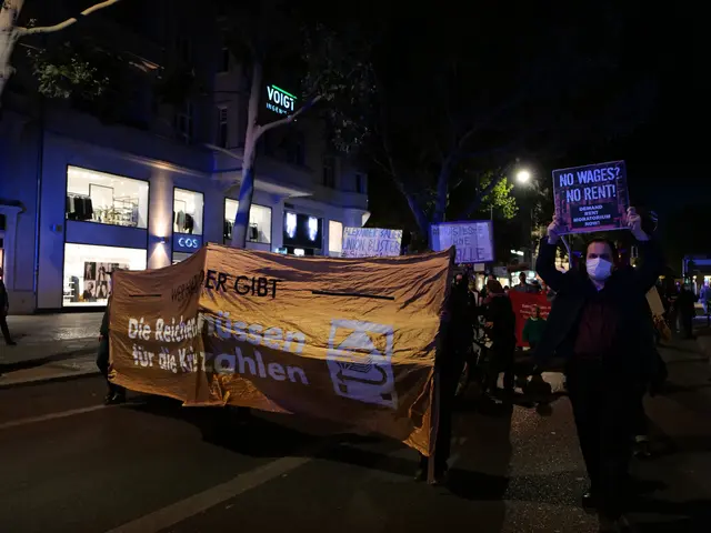 The image shows a group of people walking down a street at night, holding banners and placards with...