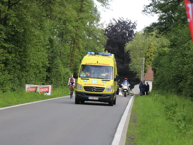 The image shows an ambulance driving down a road with a group of people riding bicycles alongside...