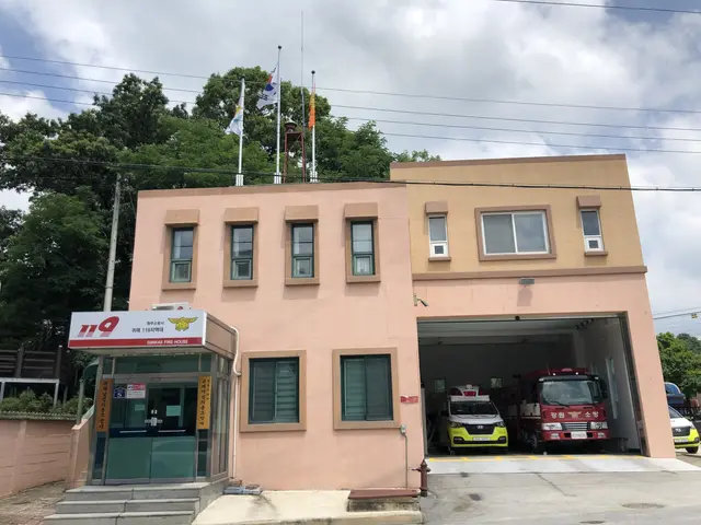 The image shows a fire station with two fire trucks parked in front of it. The building has windows...