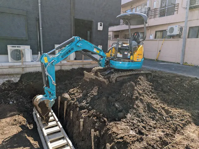 The image shows an excavator digging a hole in the ground with a ladder, surrounded by buildings...