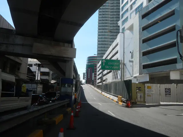 The image shows a city street with traffic cones on the side of it, vehicles on the road, a bridge...