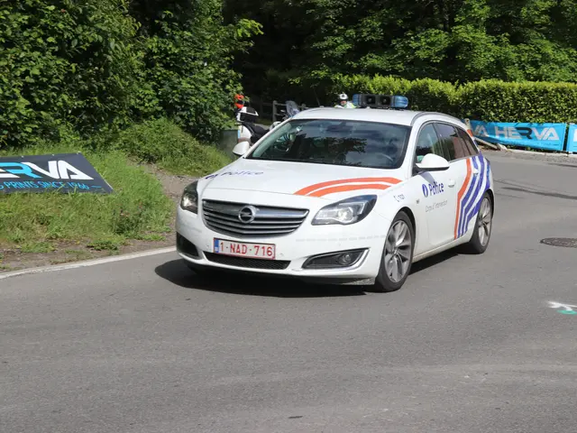 The image shows a police car driving down a road next to a forest. There are people standing on the...