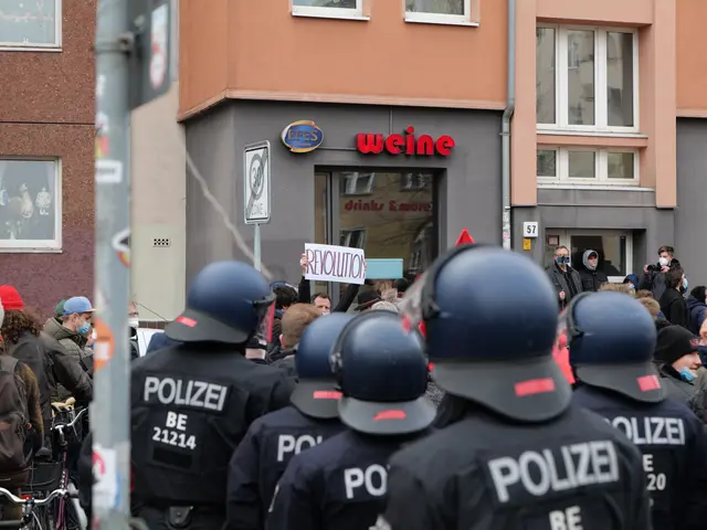 The image shows a group of police officers standing in front of a crowd of people, some of whom are...