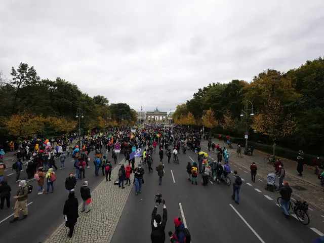 The image shows a large group of people walking down a street lined with trees and light poles,...