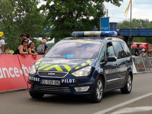The image shows a police car driving down a street next to a crowd of people, some of whom are...