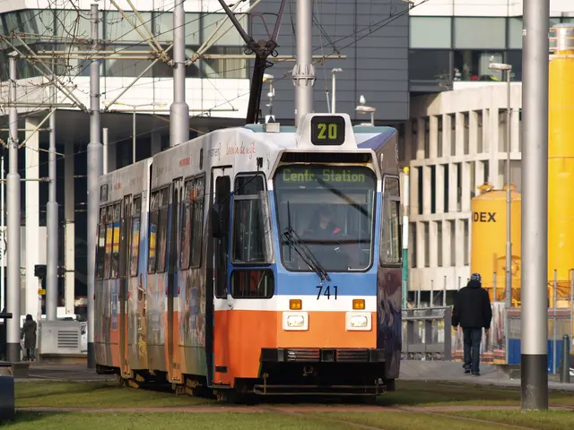 The image shows a blue and orange tram on a city street, with a person inside the train. On the...