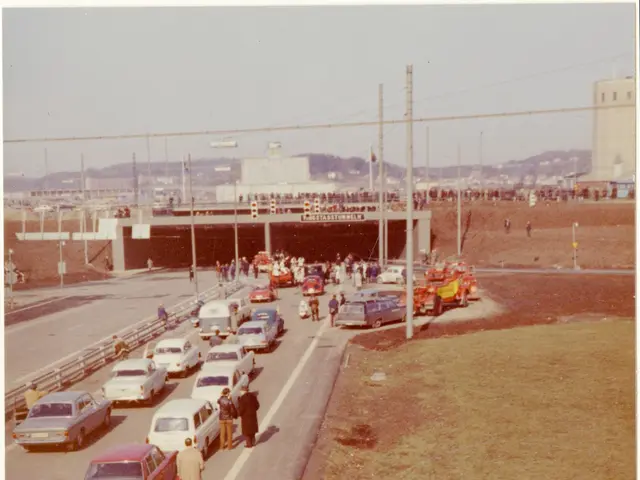 The image shows an old photo of a busy highway with cars and people on it. We can see vehicles on...
