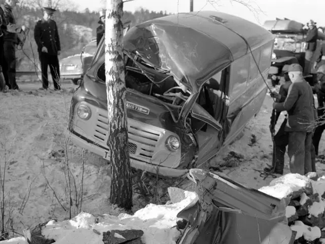 The image shows a black and white scene of a car that has crashed into a tree in the snow. There...