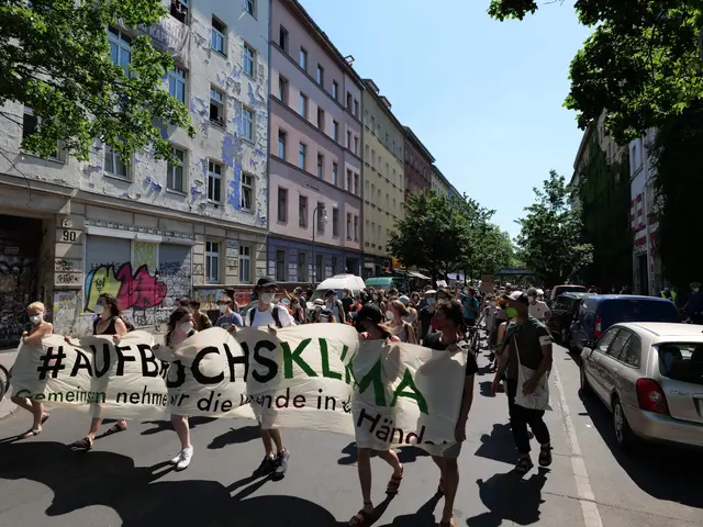 The image shows a group of people walking down a street, holding a banner with text on it. On the...