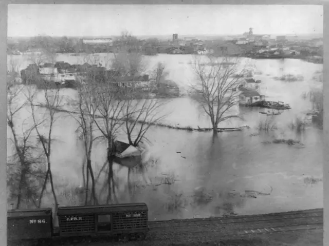 The image shows a black and white photo of a train traveling through a flooded city, with trees in...