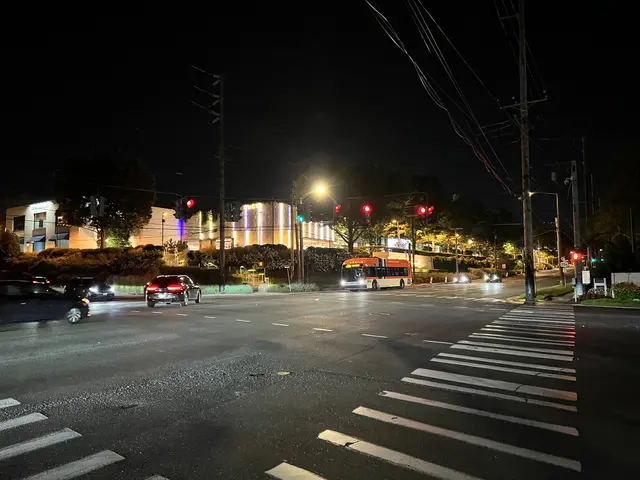 The image shows a city street at night with cars and a bus driving down it. There are street...