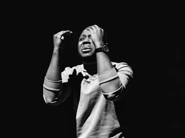 The image shows a man standing in front of a black background with his hands on his head, wearing a...