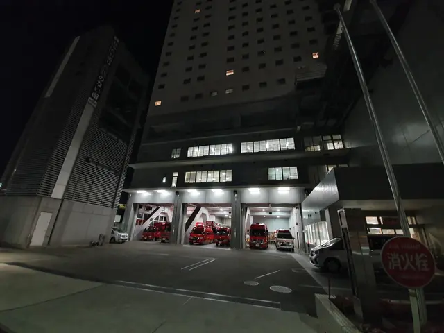 The image shows a fire station at night with several fire trucks parked in front of it. The...