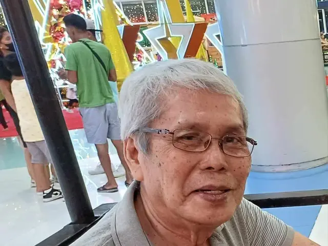 The image shows an older man wearing glasses and a watch, leaning against a railing in front of a...