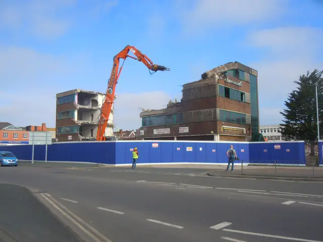 The image shows a construction site with a large building being demolished, surrounded by a fence,...
