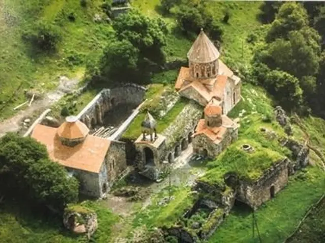 The image shows an aerial view of a small church nestled in the middle of a lush green hillside,...