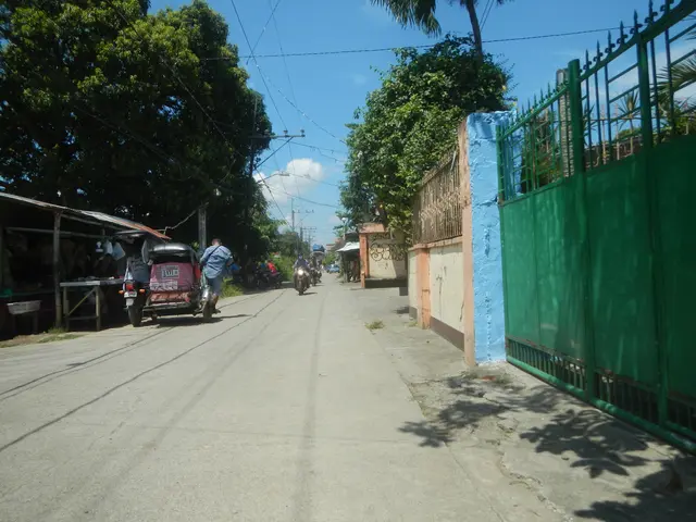 The image shows a street with people riding motorcycles down it, a green fence on the right side,...