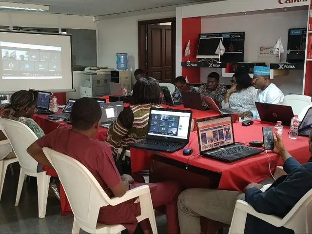 The image shows a group of people sitting around a table with laptops, mouses, bottles, and other...