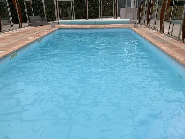 The image shows a large indoor swimming pool in a large building with glass walls, surrounded by...