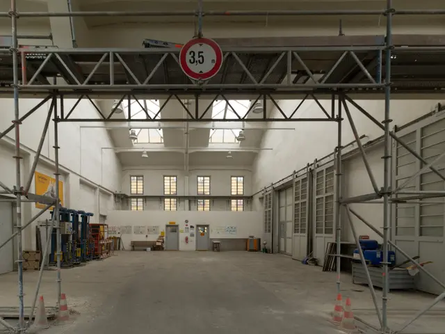 The image shows a large warehouse with a lot of scaffolding around it. There are traffic cones on...