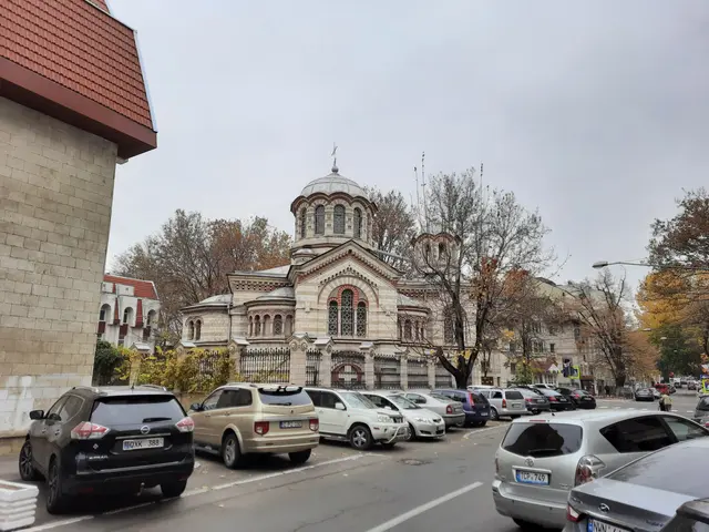 The image shows a street with cars parked on the side of it, buildings with windows, trees, light...