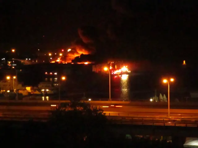 The image shows a large fire burning in the middle of a city at night, with street lights...