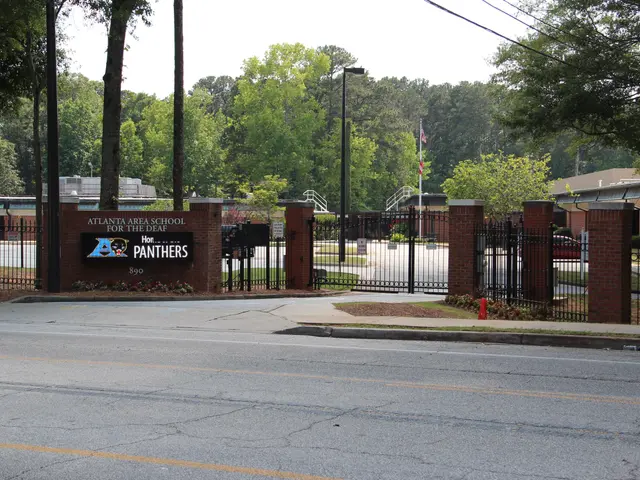 The image shows the entrance to a school with a sign on the side of it, surrounded by trees, street...
