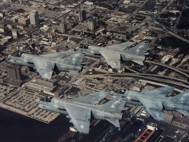 The image shows an aerial view of four fighter jets flying over a city, with buildings, roads,...