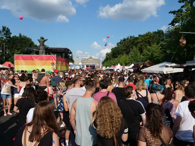 The image shows a large crowd of people walking down a street lined with tents, trees, poles,...