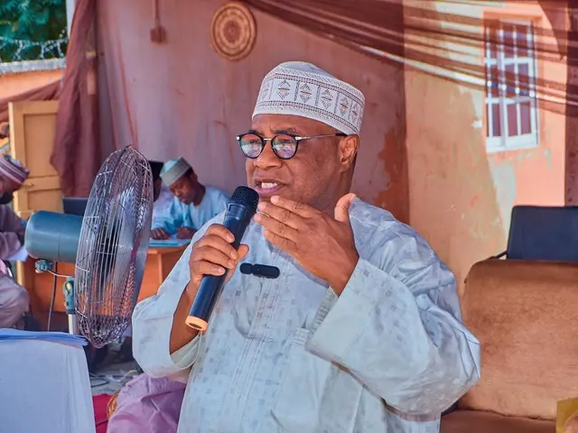 The image shows a man wearing a cap and spectacles, holding a microphone in front of a crowd of...