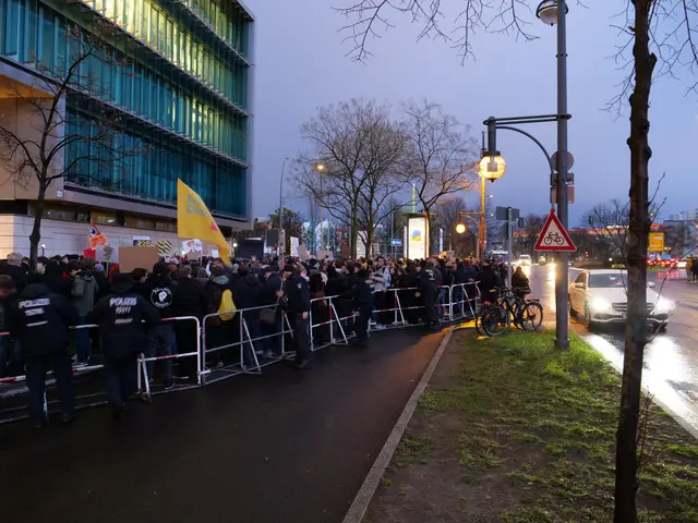 The image shows a large group of people standing in front of a building, with barricades, bicycles,...