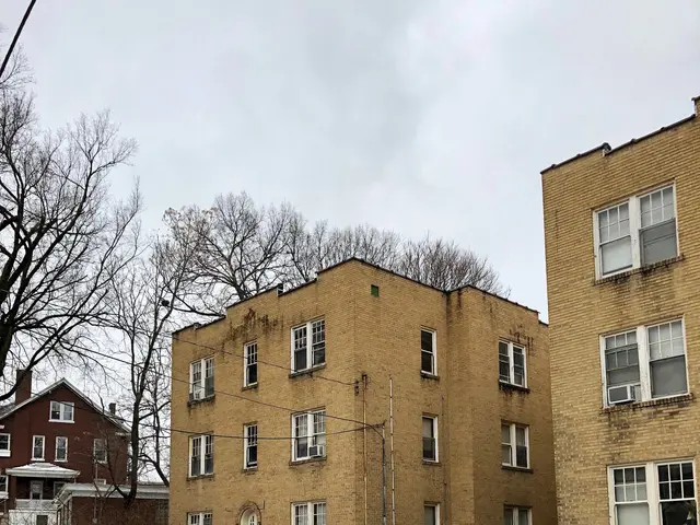 The image shows an apartment building with snow on the ground in front of it, surrounded by grass,...