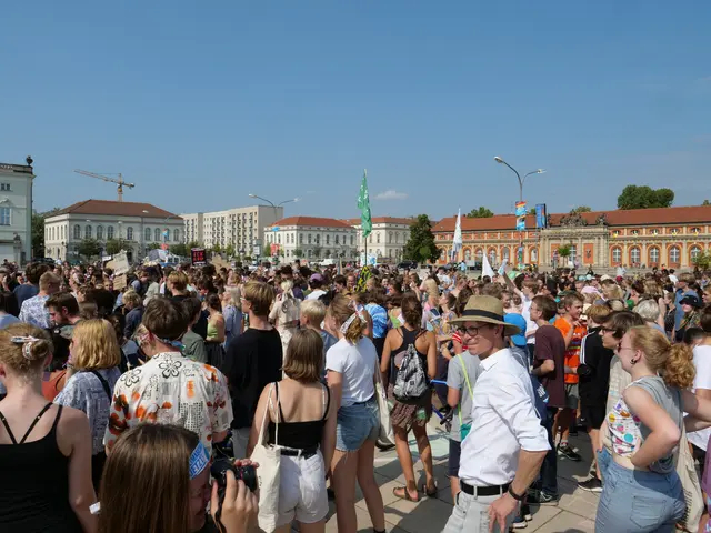 The image shows a large crowd of people standing in front of a building with windows, light poles,...