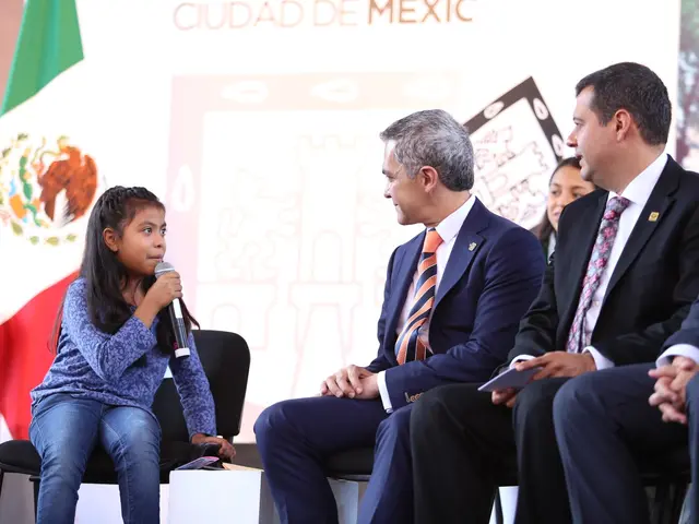 The image shows a group of people sitting on chairs in front of a Mexican flag. The girl in the...