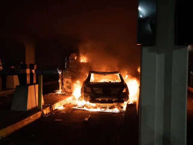 The image shows a car engulfed in flames on the side of a road at night, with a pillar on the right...