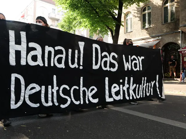 The image shows a group of people walking down a street, holding a black banner with the words...