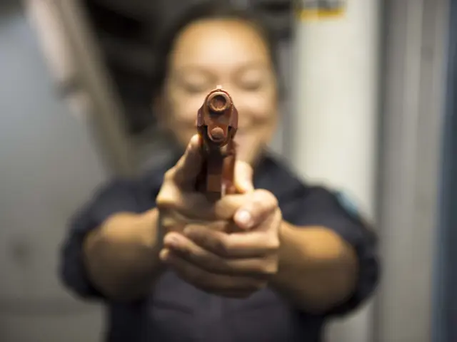 The image shows a woman holding a handgun in her hand, with a blurred background. She appears to be...
