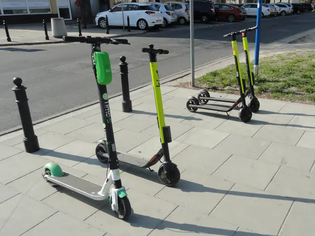The image shows three electric scooters parked on the side of a street, surrounded by grass, poles,...