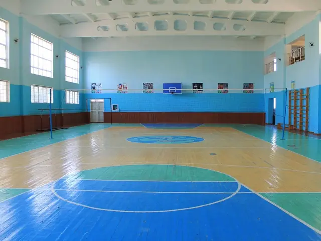 The image shows a large gymnasium with a blue and white floor, a basketball court, a net, poles, a...