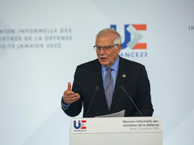 The image shows a man in a suit and tie giving a speech in front of a podium with two microphones...
