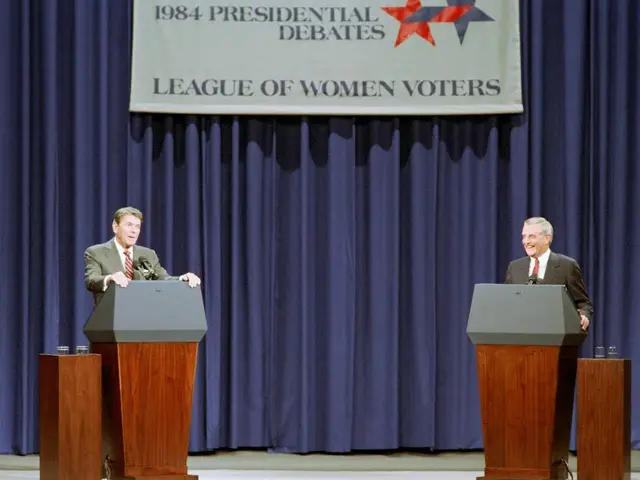 The image shows two men standing at podiums in front of a blue curtain, both wearing blazers, ties,...