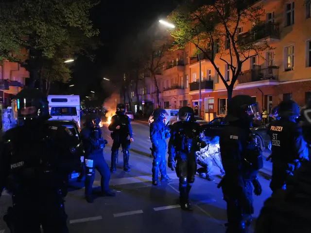 The image shows a group of police officers in riot gear walking down a street at night. They are...