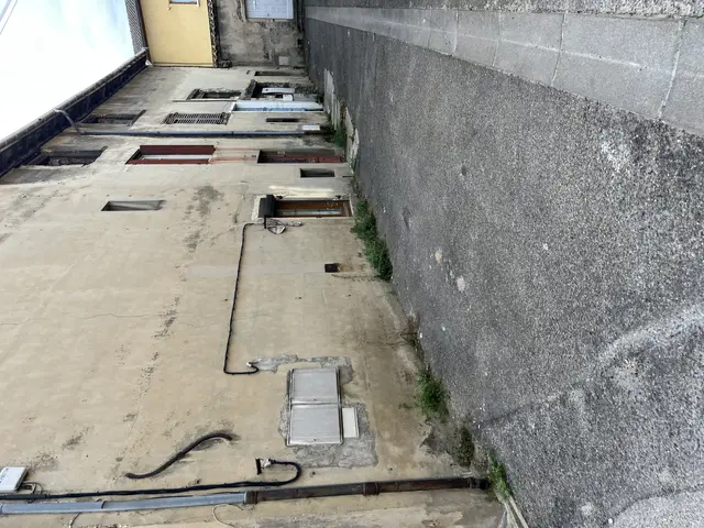 The image shows a building with a lot of pipes on the side of it, a wall on the right side, and a...