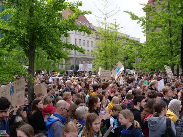 The image shows a large crowd of people standing in front of a building, holding placards with text...