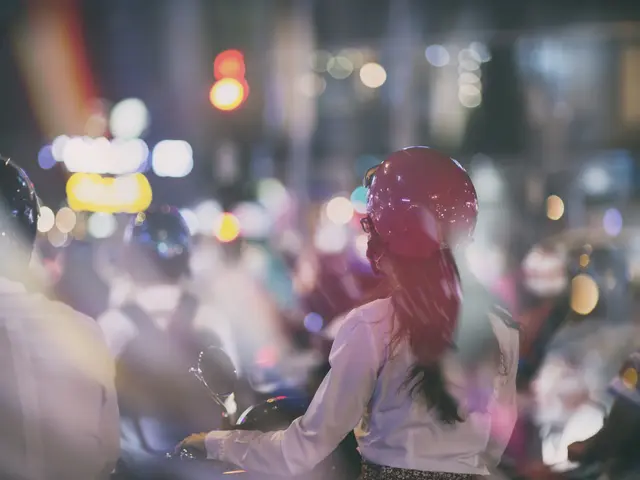 The image shows a woman riding a motorcycle down a busy city street at night. She is wearing a...