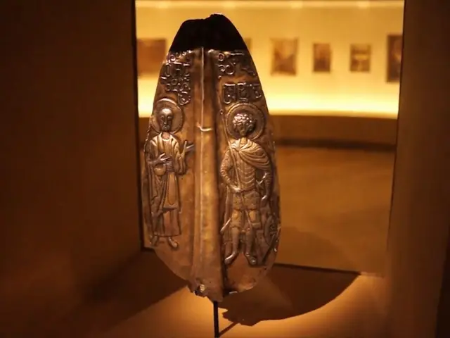 The image shows a metal object in a glass case on display in a museum, with a wall of photo frames...
